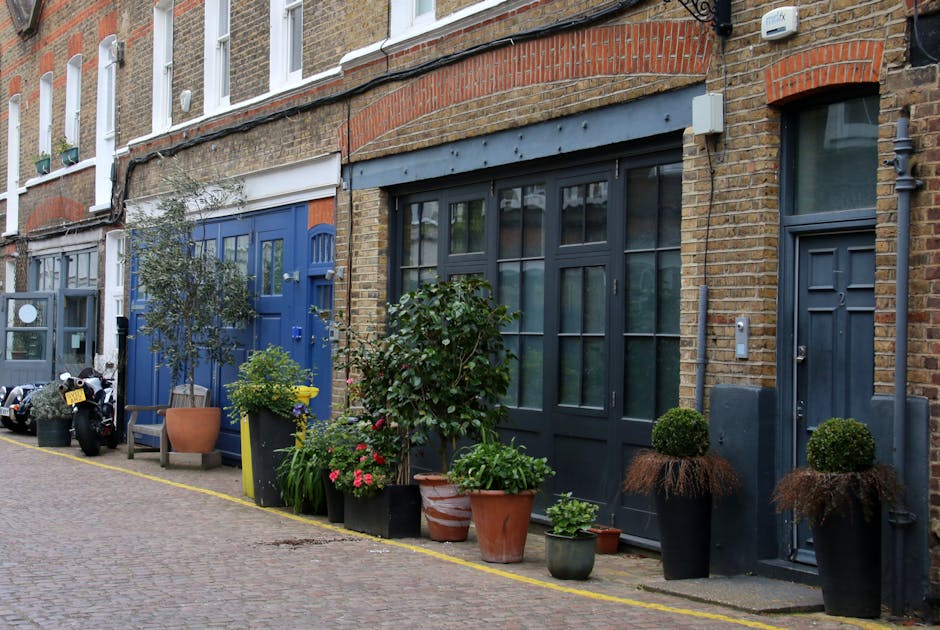 A narrow cobblestone alleyway between white terraced buildings with black wrought iron balconies and window frames, featuring a variety of potted plants and small trees along the sides. On the left, t